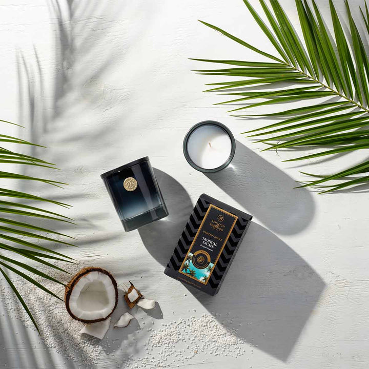 Tropical Escape Scented Candle on a wooden surface surrounded by coconut and palm ferns