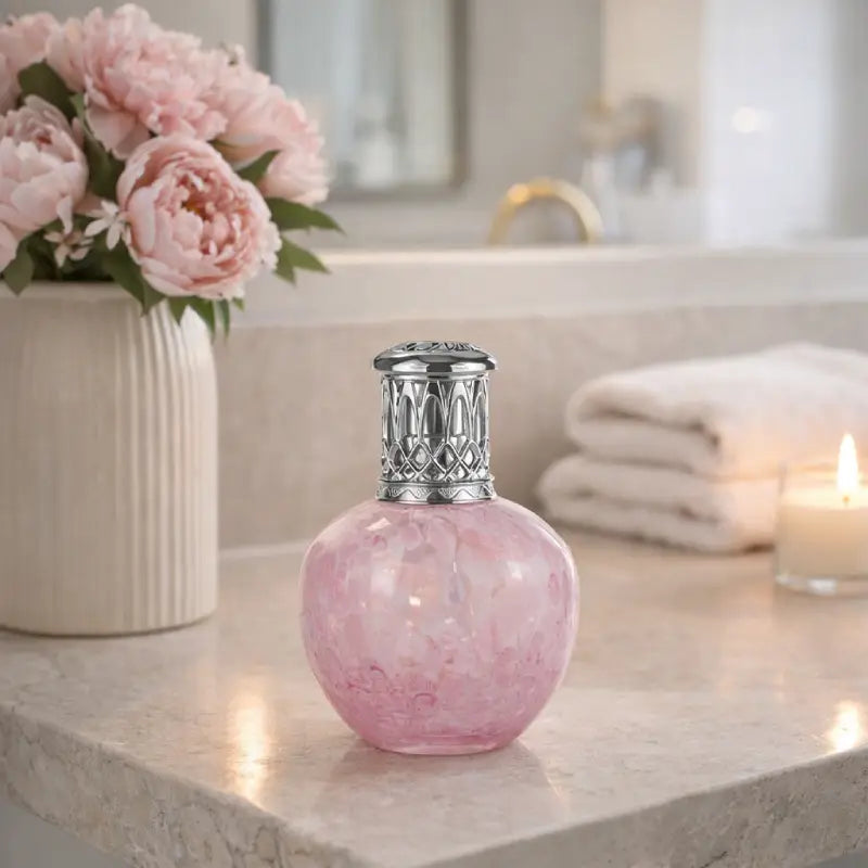 Pink marble-textured bottle with silver cap on a bathroom counter, next to a vase of pink flowers and a lit candle.