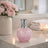 Pink marble-textured bottle with silver cap on a bathroom counter, next to a vase of pink flowers and a lit candle.