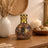 Decorative lamp with mosaic design on a wooden surface, surrounded by a vase with dried plants and a candle.